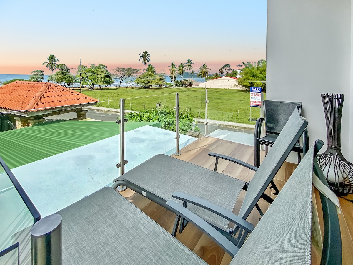 A front patio is furnished with sleek lounge chairs positioned for optimal ocean views. The scene captures vibrant greenery and a sandy beach beyond, with an expansive sky tinged with soft pastel colors as the sun sets on the horizon.