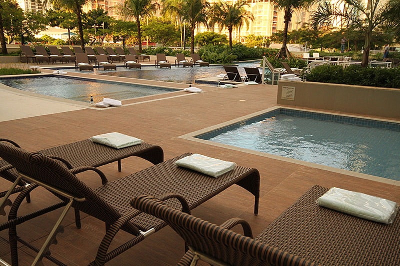 Lounge chairs with light-colored cushions are positioned around a spacious pool area that features multiple pools, including a children's pool. Lush greenery and palm trees are visible in the background, creating a serene outdoor space enhanced by natural light.