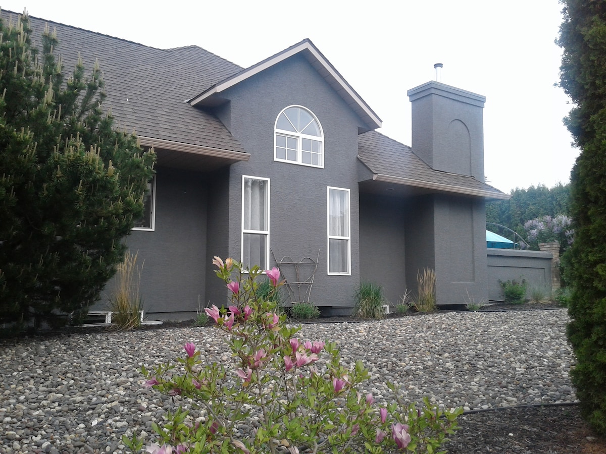 The exterior of the home features a grey facade with distinctive architectural elements, including a prominent front arch window and a side chimney. Lush greenery and flowering plants are visible in the landscaped yard, while gravel lines the pathway leading to the entrance.
