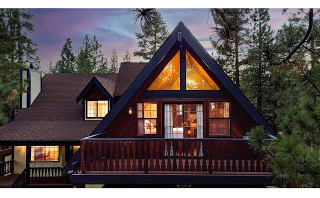 The cabin's exterior showcases a charming A-frame structure with warm, glowing windows that illuminate the upper level. Surrounded by tall trees, a balcony extends from the front, bordered by a wooden railing, inviting relaxation amidst nature.