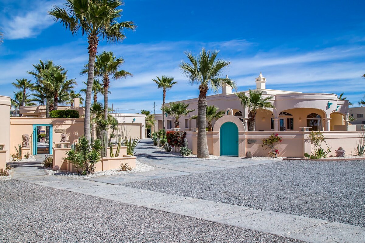 The exterior features a well-maintained courtyard with palm trees and desert landscaping. A light-colored building with arches and a distinctive turquoise door can be seen, set against a clear blue sky. A gravel pathway leads to the entrance.