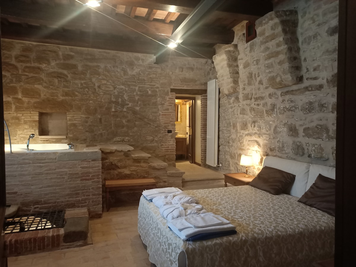 The room features exposed stone walls and wooden beams, creating a rustic ambiance. A spacious bed with a patterned coverlet occupies the center, flanked by wooden nightstands with lamps. An elegant soaking tub is visible, along with an entrance leading to the bathroom.