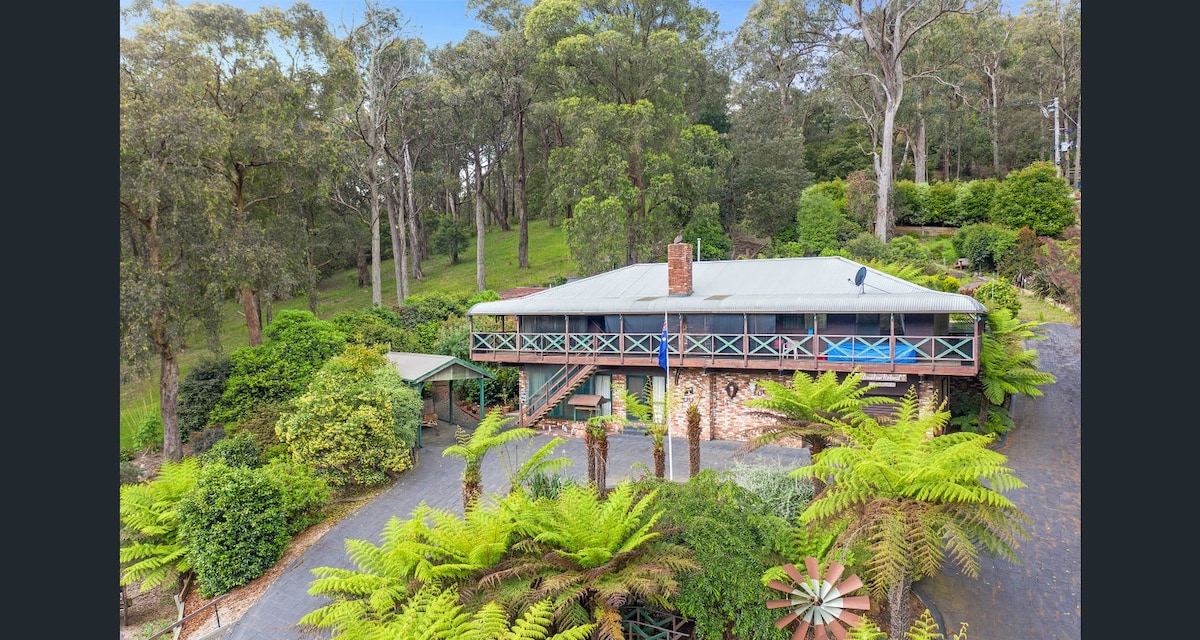 The two-level home is surrounded by lush greenery and tall trees, offering a tranquil setting. A spacious deck can be seen at the front, providing outdoor relaxation space. The structure features natural materials and blends harmoniously with its serene environment.