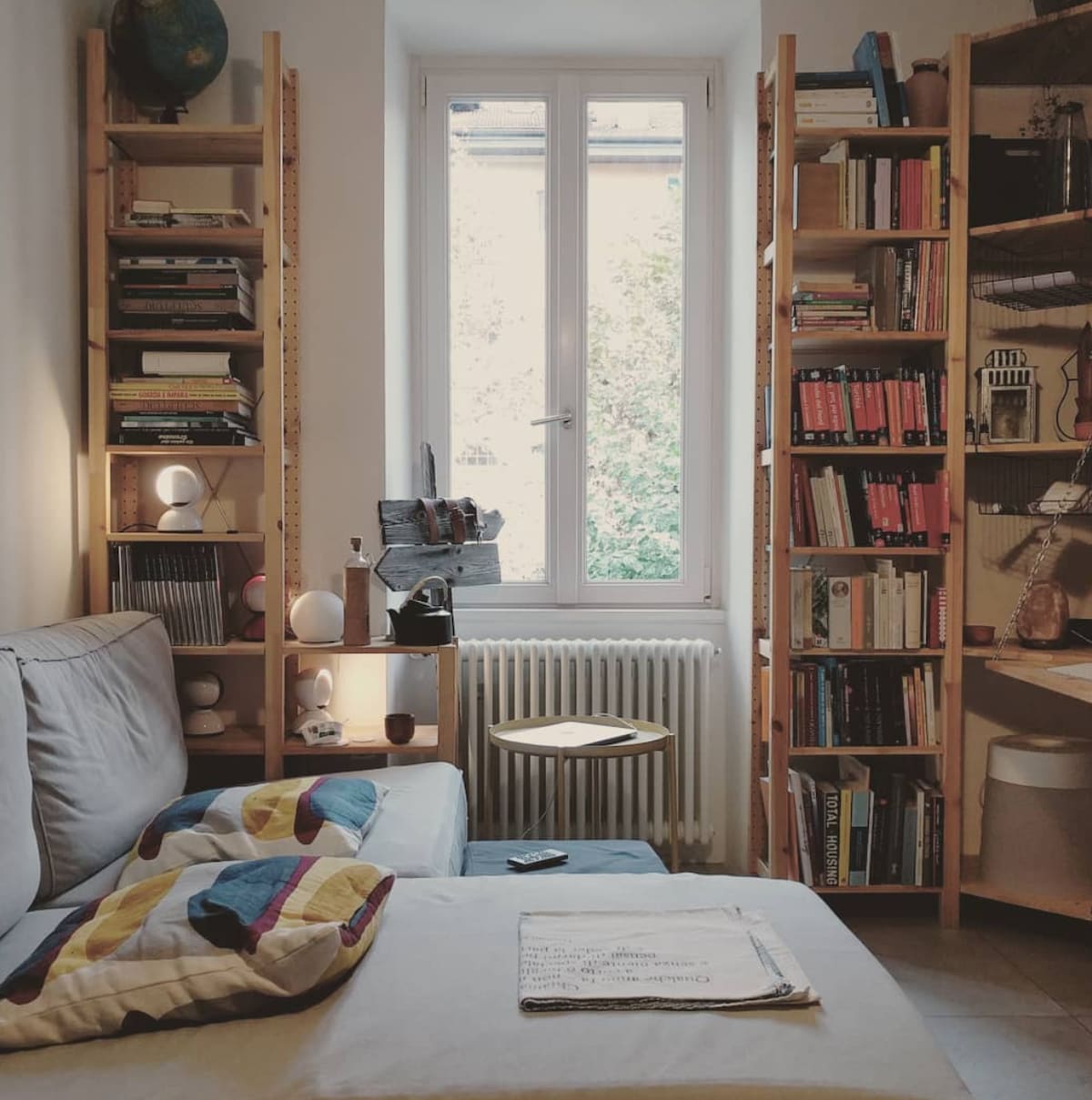 A cozy seating area is presented with a comfortable sofa adorned with colorful cushions. Two tall bookshelves filled with books line the walls. Natural light enters through a window, illuminating a small side table next to the sofa. A warm atmosphere is created by soft lighting.