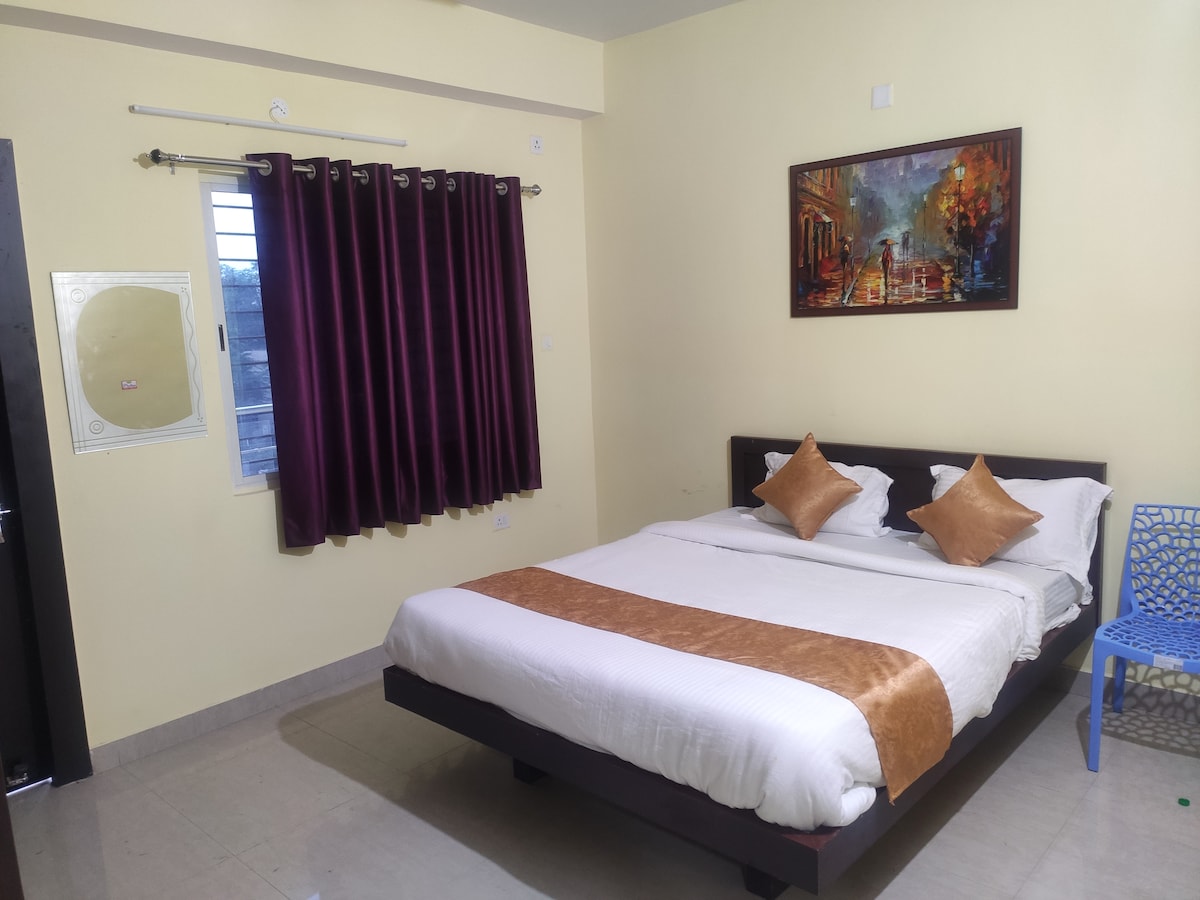 A well-lit sleeping area features a spacious bed with two decorative cushions. Rich purple curtains frame a window, and a colorful painting adorns the wall. A blue chair adds a pop of color, while the light tile floor enhances the room's overall brightness.