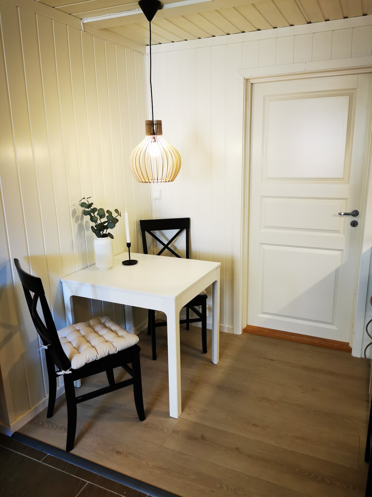 A small dining space features a white table complemented by two black chairs. A soft, stylish pendant light illuminates the area, while a candle and a small plant sit on the table. The walls are lined with light-colored wood paneling.