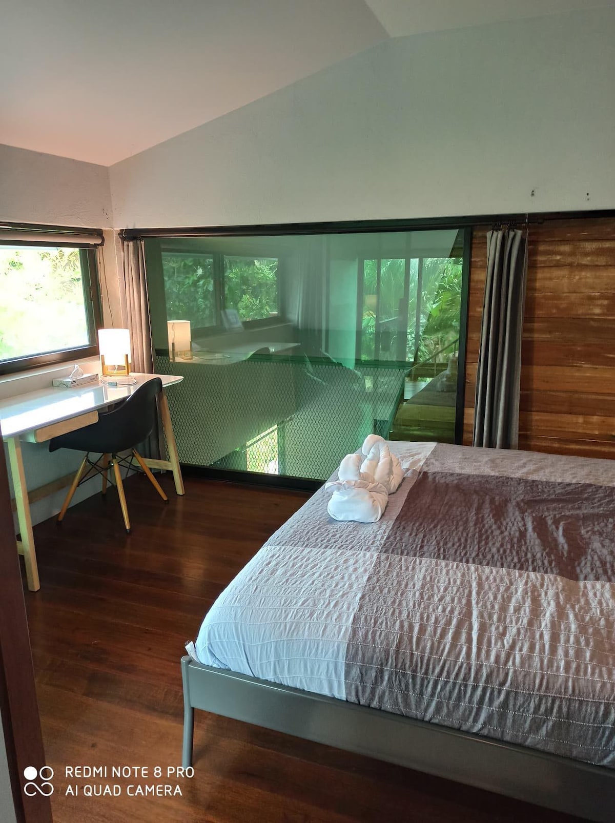 A serene bedroom is presented with a spacious bed covered in a light gray quilt. A small wooden desk with a chair is positioned by a large glass window overlooking greenery. Soft lighting is provided by a nearby lamp, enhancing the tranquil ambiance.