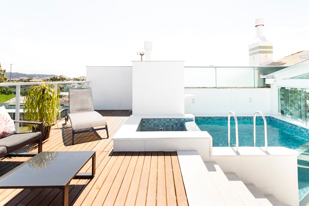 An outdoor terrace features a private pool alongside a seating area. Sun loungers and a glass table set atop a wooden deck provide relaxation options. A modern design and surrounding greenery create a serene atmosphere, complemented by ample natural light.