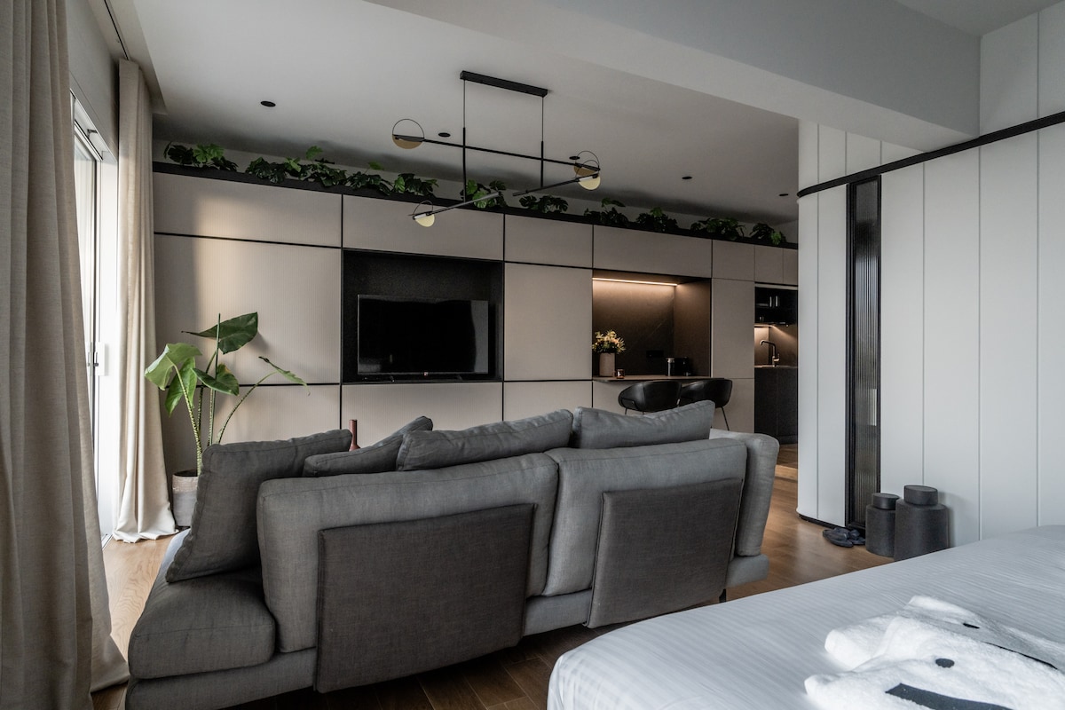 A modern living area features a spacious gray sofa, providing ample seating. Above, greenery adorns the ledge, enhancing the room’s natural feel. A flat-screen television is mounted on the wall, while a kitchenette is visible in the background, offering convenience for guests.