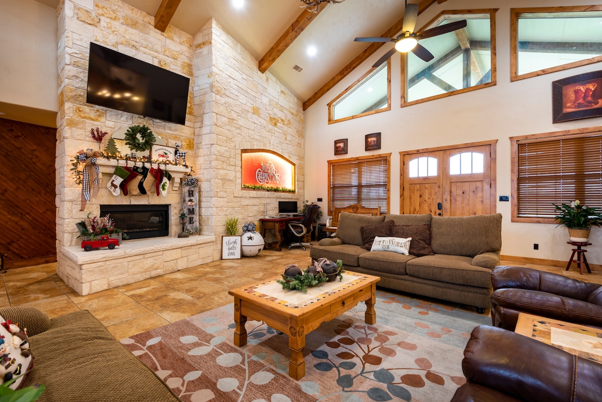 A spacious living area features gleaming stone walls and vaulted ceilings with wooden beams. Comfortable seating arrangements are present, including a large sectional sofa and leather chairs. A media unit and decorative elements add visual interest, while natural light filters through the large windows.