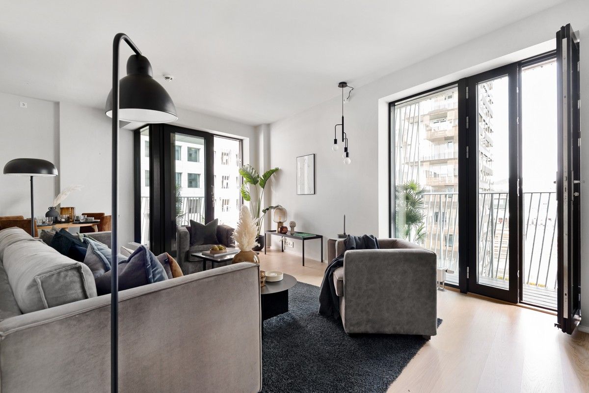 A spacious living area features a comfortable sectional sofa adorned with decorative pillows. Large glass doors open to a bright balcony, allowing natural light to fill the room. A sleek floor lamp and coffee table complement the modern aesthetic, while a rug adds a cozy touch.