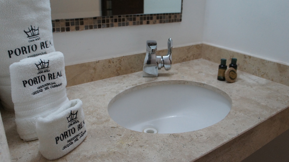 A clean bathroom space is features a round sink set into a polished stone countertop. Neatly arranged towels are displayed beside the sink, accompanied by small bottles of toiletries. A mirror with a decorative border adds depth to the room.