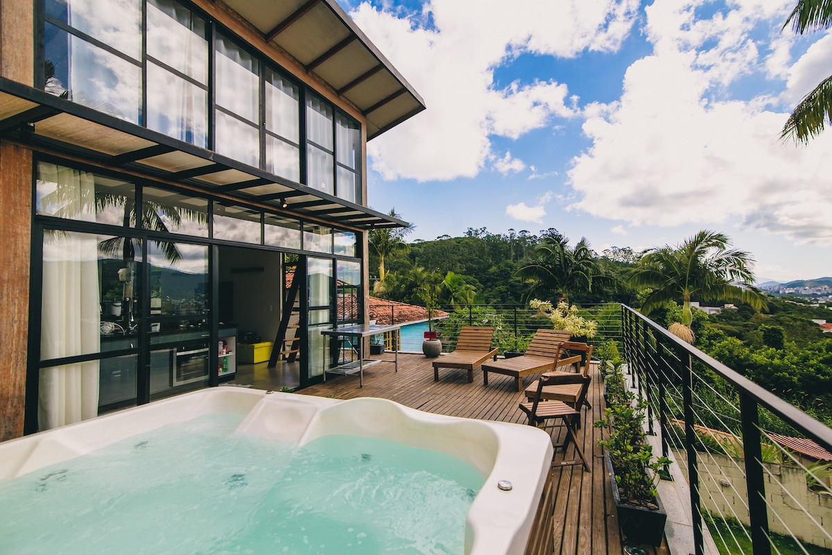 A modern house is presented with large glass windows and an inviting deck area. A bubbly jacuzzi is featured alongside several lounge chairs, all set against a backdrop of lush greenery and a clear sky with scattered clouds.