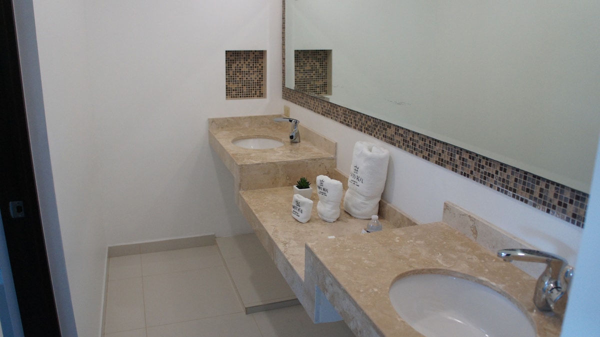 The bathroom features dual sinks set in a neutral-toned countertop with a mosaic tile accent above. A large mirror reflects the tidy space, complemented by subtle decorative elements and a light color scheme for a clean and modern feel.