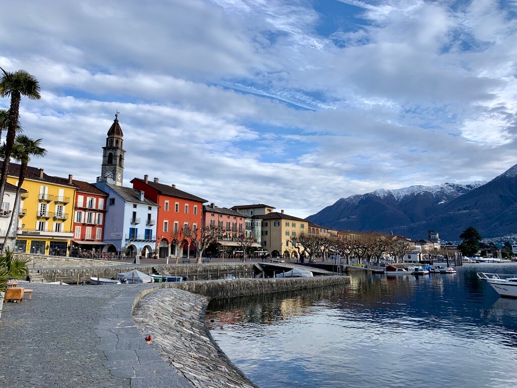 A serene waterfront scene is captured, showcasing colorful buildings lining the shore. A pathway runs alongside the calm water, reflecting the sky and surroundings. Snow-capped mountains loom in the background, providing a scenic backdrop to the tranquil atmosphere.