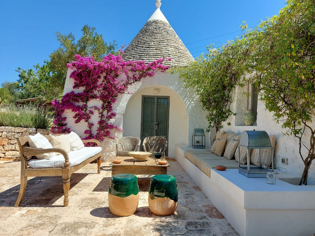 A charming outdoor seating area is framed by vibrant pink bougainvillea. Natural wood furniture, including a sofa and two green-accented side tables, creates a warm space. A built-in bench and various cushions provide comfort, while the white trullo structure adds to the serene environment.