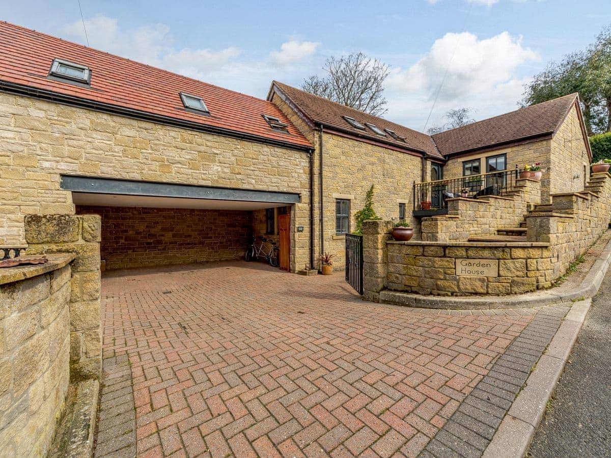 The exterior of the property features a stone facade with a welcoming entrance. A paved driveway leads to a carport, providing private parking. The surrounding area is landscaped, including steps and a small patio area, enhancing the inviting appearance of the house.