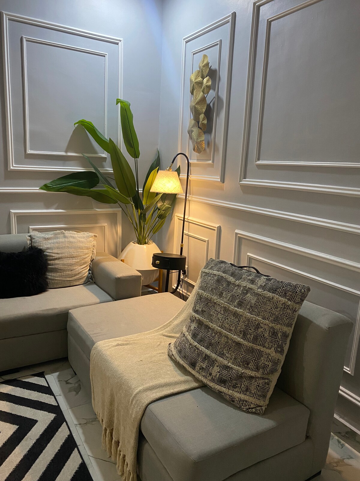 A corner of a living area is presented, featuring a comfortable, light-colored sectional sofa adorned with various textured cushions. A throw blanket drapes casually over one section. A floor lamp provides soft light, complemented by a tall, green plant against a textured wall.