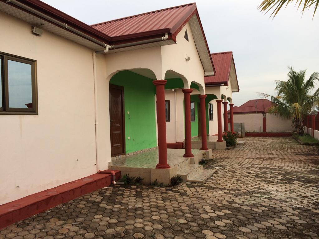A row of standard double rooms is configured with smooth, colorful façade accents. Each space features a contrasting green and neutral exterior, supported by red pillars. A paved pathway leads to the doors, bordered by small plants, set against a clear sky.