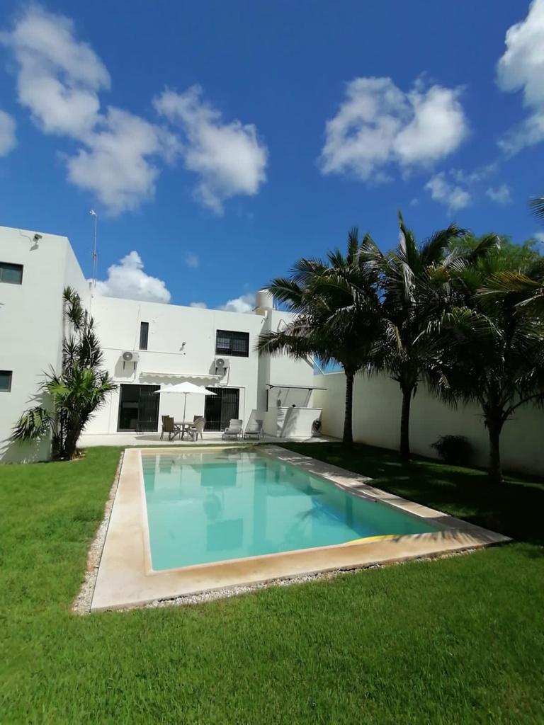 A well-maintained outdoor space features a private pool surrounded by green grass. Lush palm trees are positioned beside the pool area. The exterior of the house is visible, showcasing large windows that allow natural light into the interior.