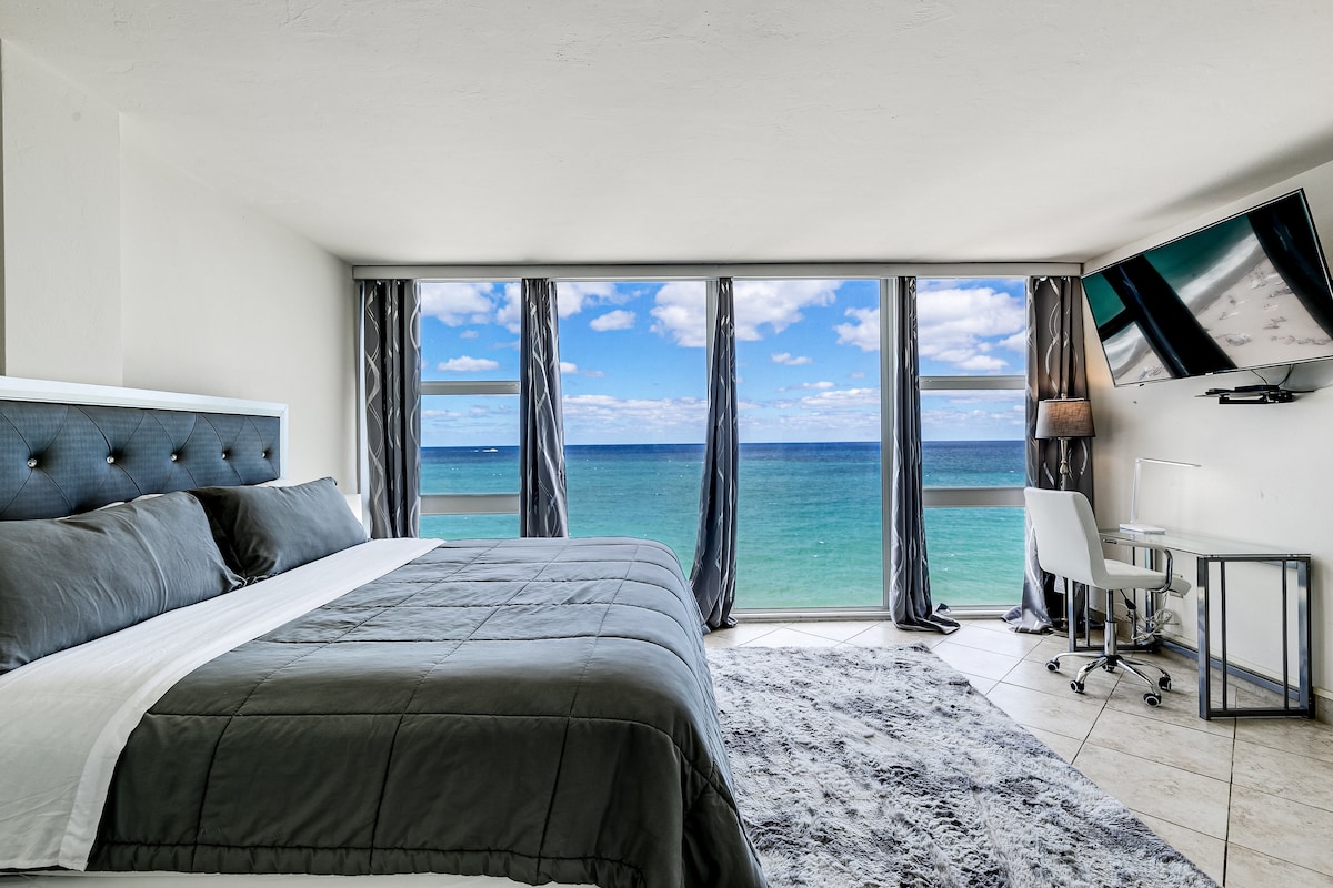 A spacious bedroom is presented with a king bed dressed in dark grey linens. Large windows offering unobstructed ocean views are flanked by sheer curtains, allowing natural light to fill the room. A desk and chair are positioned beside the windows, complementing the serene setting.