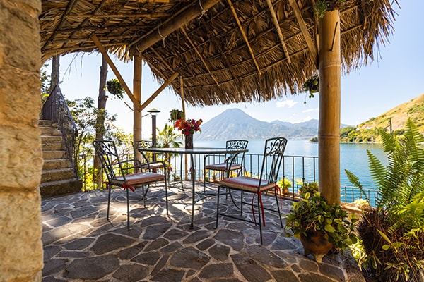 An outdoor seating area is featured under a thatched roof, offering a view of the lake and surrounding mountains. Four metal chairs surround a rectangular table, positioned on a stone patio. Lush greenery and decorative plants enhance the tranquil setting.