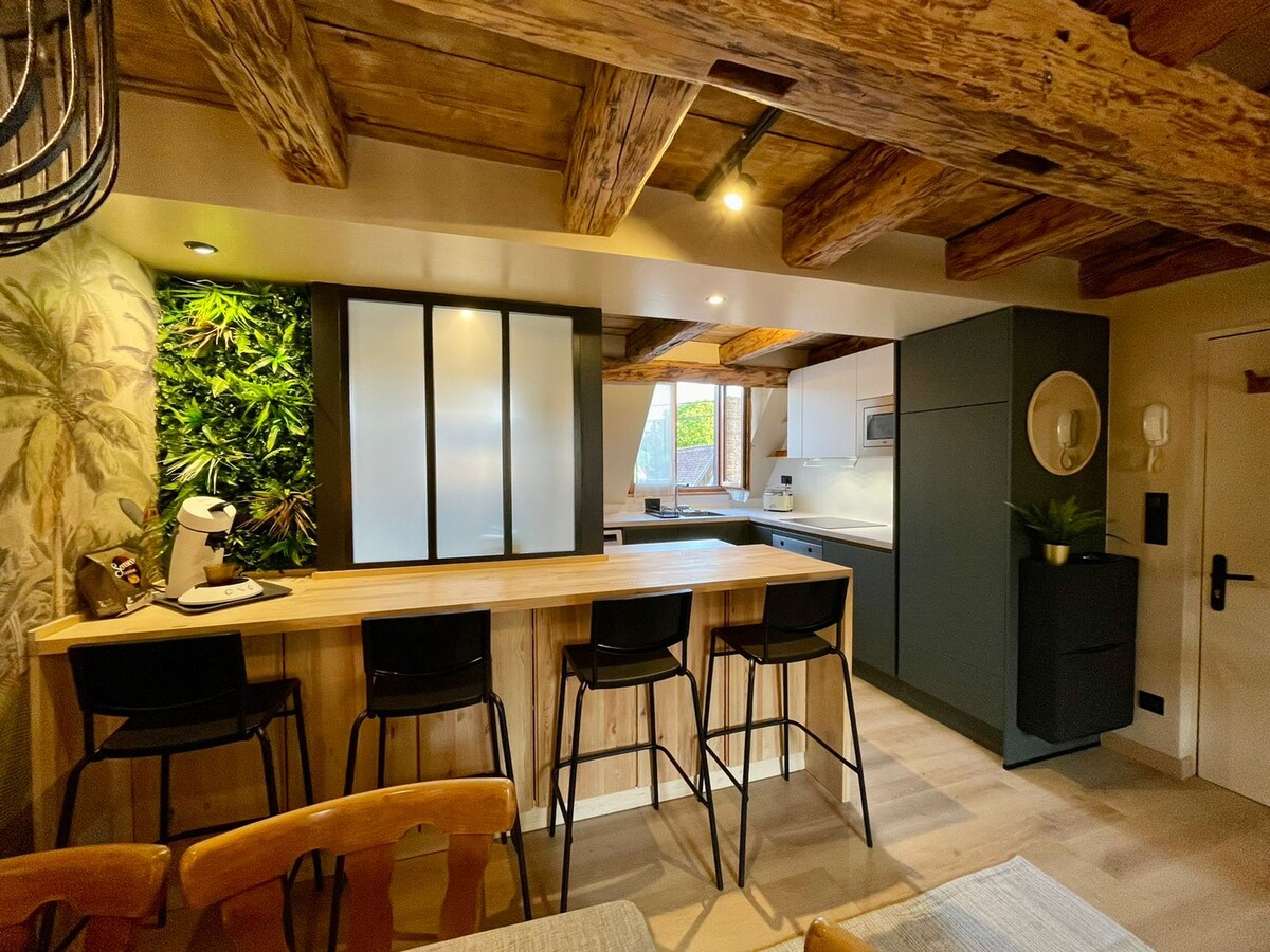 A modern kitchen area features wooden beams and a stylish bar with four black stools. Indoor plants are displayed against a textured wall, enhancing the space. A sleek countertop with a coffee machine and light cabinetry is visible, along with a window allowing natural light to enter.