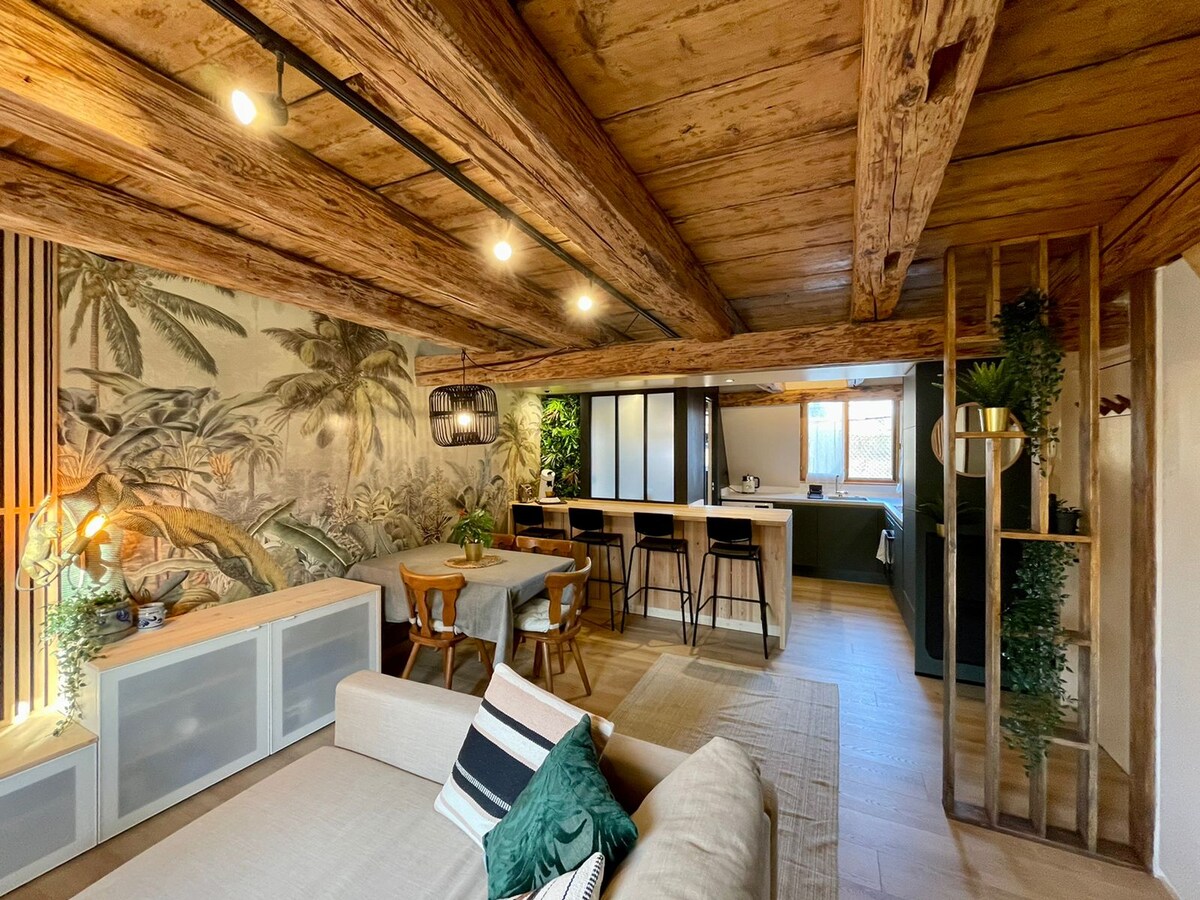 An inviting living area is enhanced by rustic wooden beams and a tropical mural. A comfortable sofa is positioned alongside a dining table for four. A modern kitchen with a long island bar complements the space, with large windows allowing natural light.