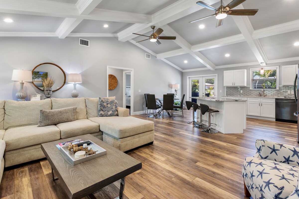 An open-concept living area showcases a spacious sectional sofa accompanied by a low coffee table. Ceiling fans are visible above, and light streams through large windows. A dining area with a modern table and chairs is adjacent to a bright kitchen featuring high-end appliances.