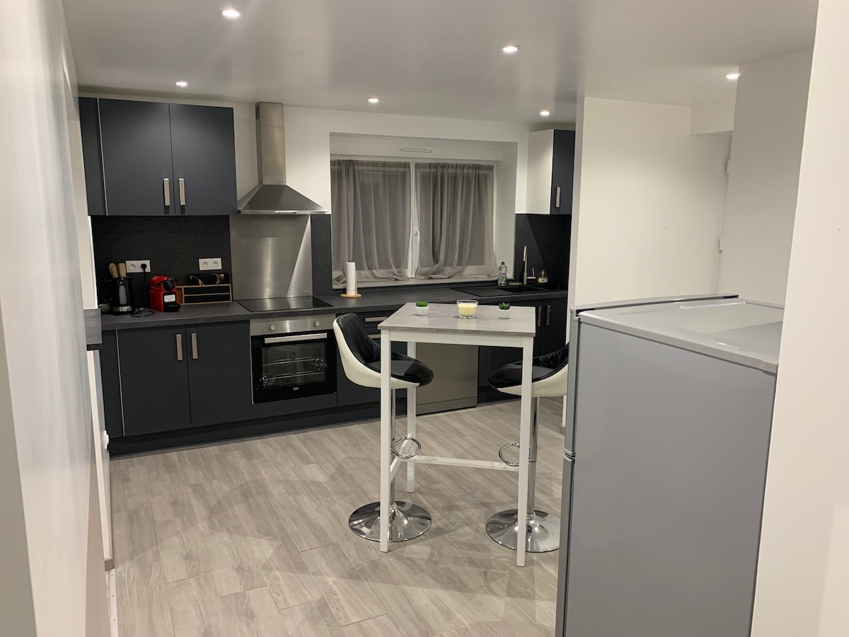 A modern kitchen features sleek dark cabinetry and stainless steel appliances. A small dining table is set with stools, providing a cozy eating space. Warm light is emitted from recessed ceiling fixtures, illuminating the grey flooring and minimalist design throughout the area.