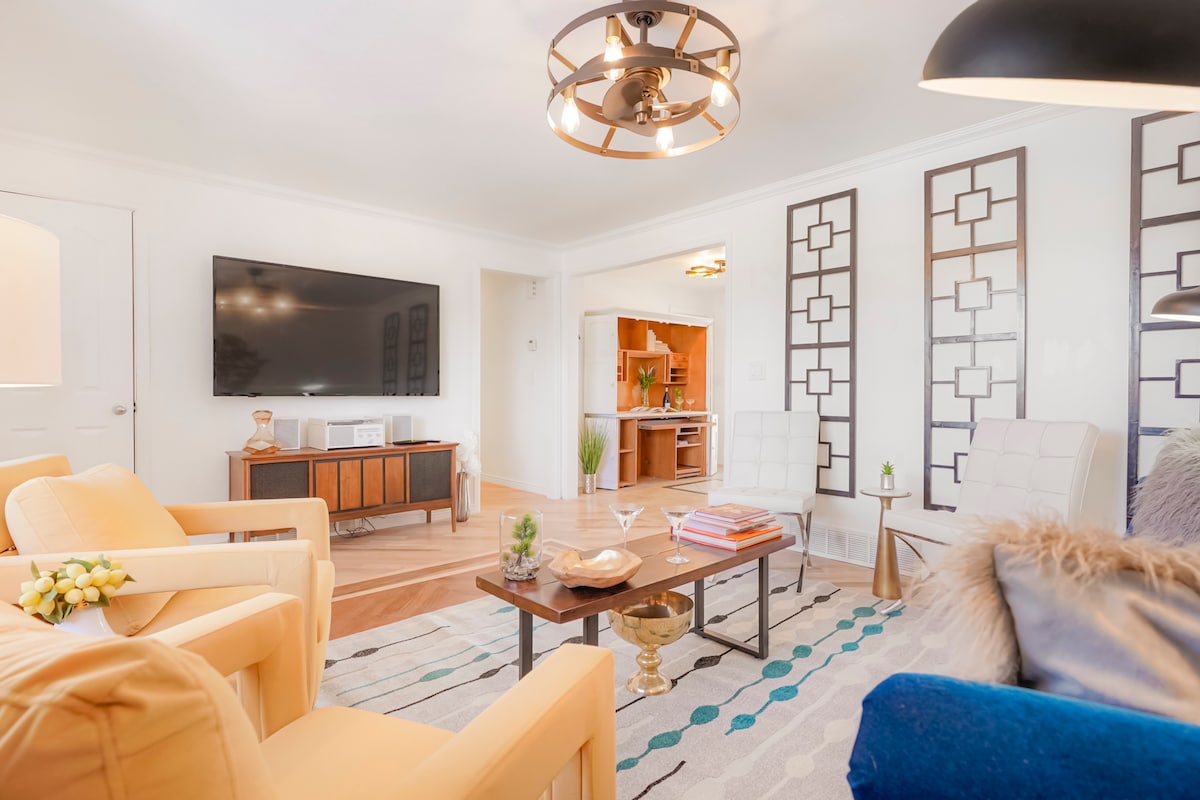 A bright living room is highlighted by a large flat-screen TV mounted on the wall. Two light-colored armchairs and a blue sofa surround a central coffee table adorned with decorative items. The space features a patterned rug and stylish light fixtures that complement the overall design.