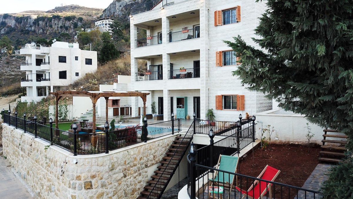 The exterior of the building is depicted, showcasing multiple stories with balcony access and orange shutters. A garden area features a pool, surrounded by a stone wall, with a wooden pergola nearby. Lounge chairs are positioned under the shade of a tree.