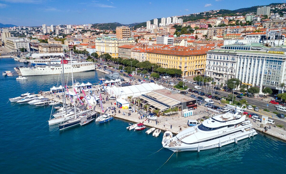 Aerial view of a lively waterfront in Rijeka, featuring numerous boats docked along a bustling marina. Colorful buildings line the shore, while a spacious promenade invites visitors to explore. Clear blue waters reflect the vibrant atmosphere of the area, with the cityscape rising in the background.