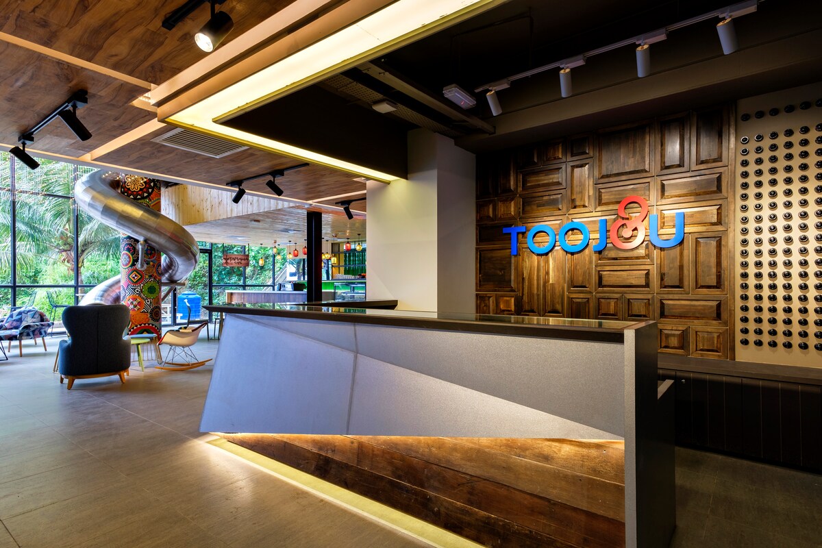 The reception area features a modern design with a sleek front desk and warm wooden accents. A colorful art installation can be seen in the background, adding a unique touch to the space. Large windows allow natural light to fill the room.