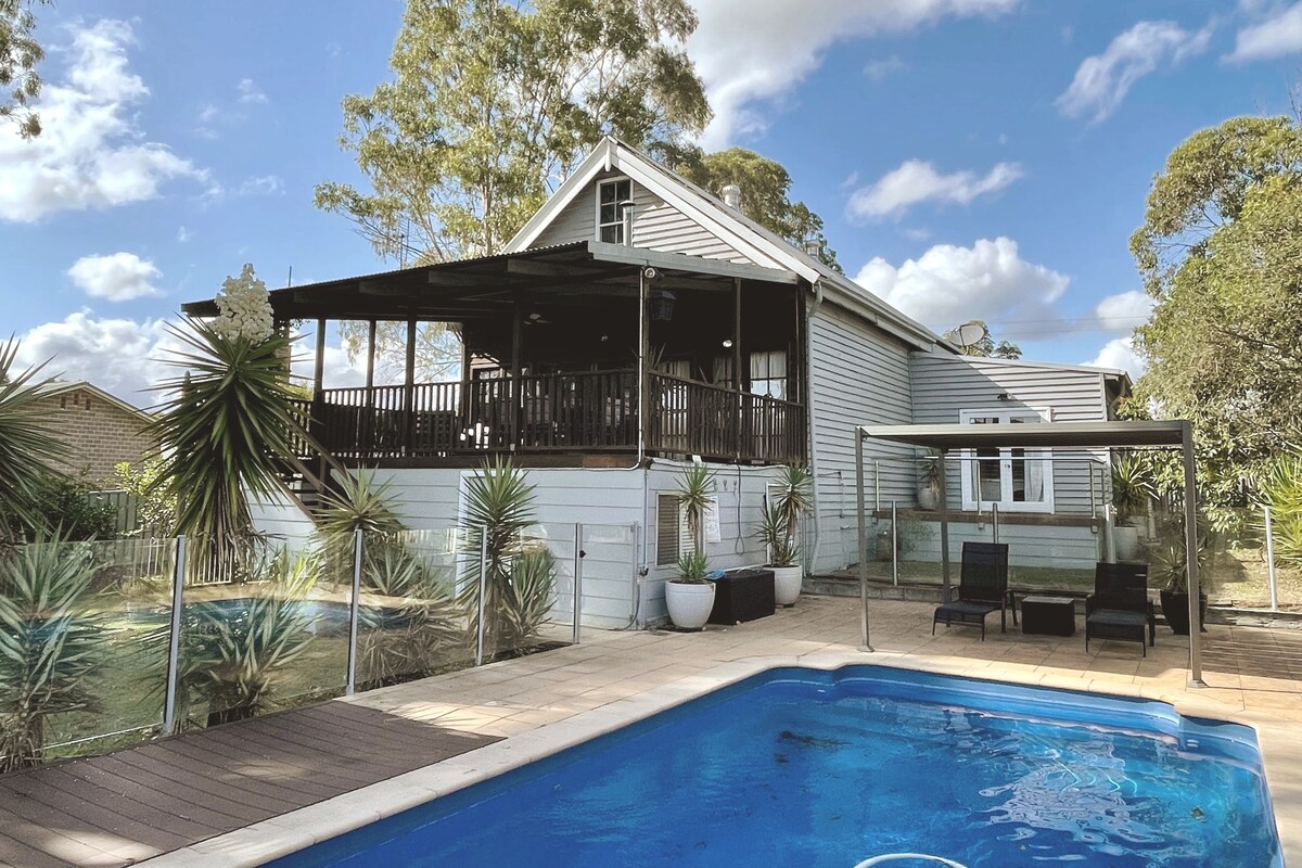 The exterior of the historic church hall conversion is showcased, featuring a spacious covered deck overlooking the backyard. A pool is visible in the foreground, framed by light-colored tiles and surrounded by plants, while the architecture reflects a charming rustic style.