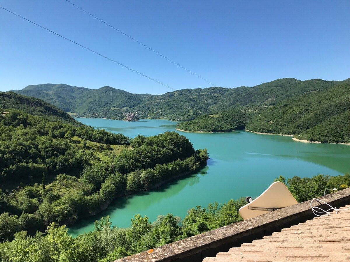 A panoramic view showcases a serene lake surrounded by lush green mountains. The water's surface reflects vibrant shades of turquoise, while the landscape features a mix of trees and hills, creating a peaceful natural setting.