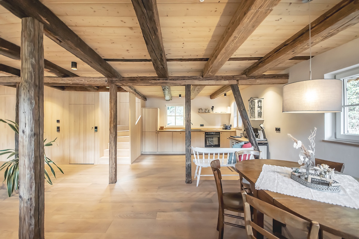 A spacious living area is framed by sturdy wooden beams, enhancing the rustic charm. A round dining table with wooden chairs occupies one corner, while a modern kitchen featuring light wood cabinetry is visible in the background. Natural light floods the space, highlighting the warm tones of the wood flooring.