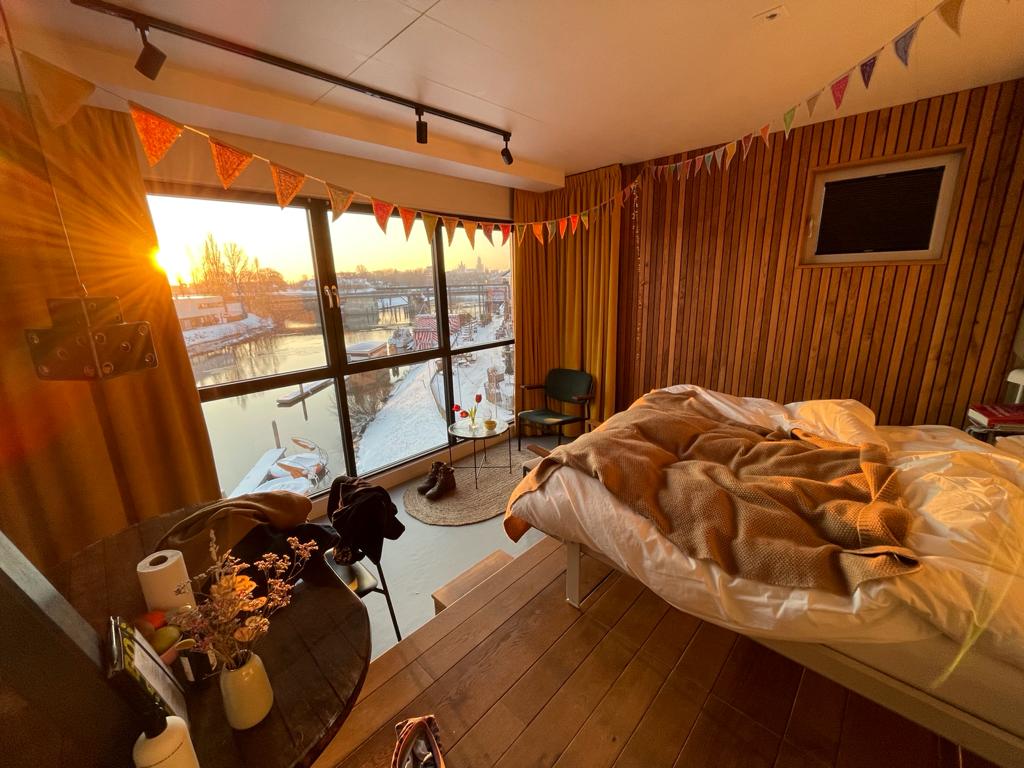 A cozy hotel room features warm wooden walls and large windows showcasing a sunset over the water. A spacious bed adorned with soft bedding is complemented by a small dining table with decorative items. A comfortable chair occupies one corner, adding to the inviting ambiance.