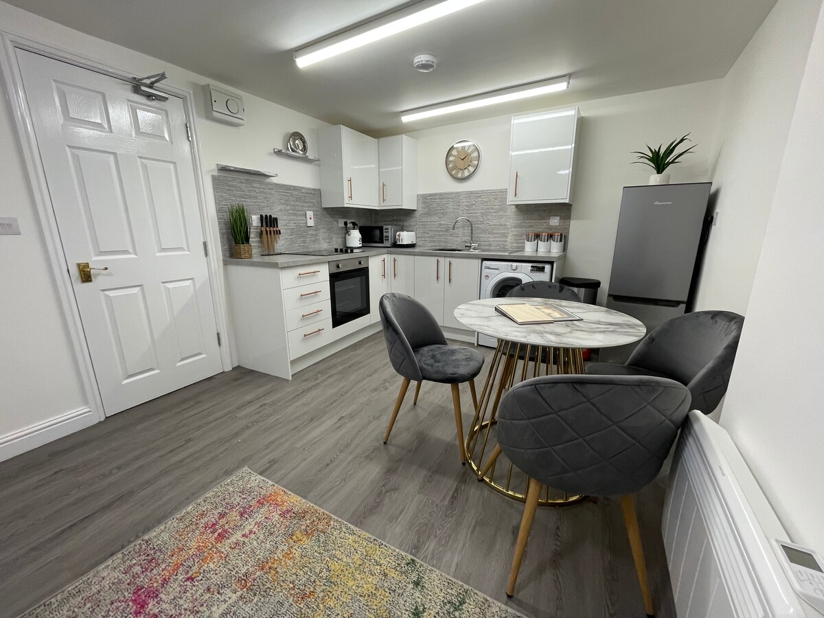 A modern kitchen area features sleek white cabinetry and a stylish marble-top dining table surrounded by four upholstered chairs. A refrigerator and various kitchen appliances are visible, with contemporary décor adding a touch of warmth to the space.