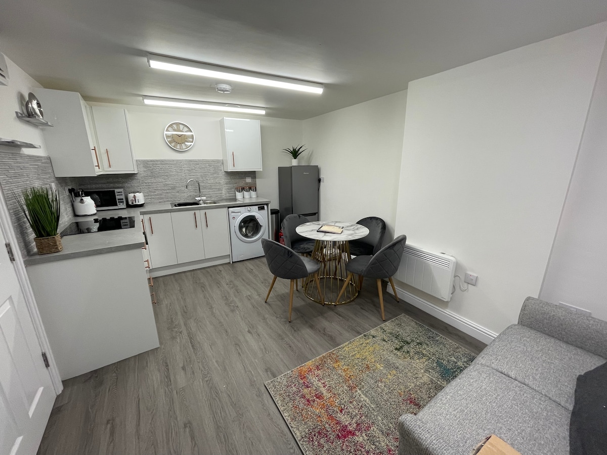 A modern kitchen area is complemented by white cabinetry and sleek gray countertops. A round dining table with gold legs is surrounded by two gray chairs. A washing machine and refrigerator are positioned against the wall, while a cozy seating area features a soft, gray sofa and a colorful area rug.