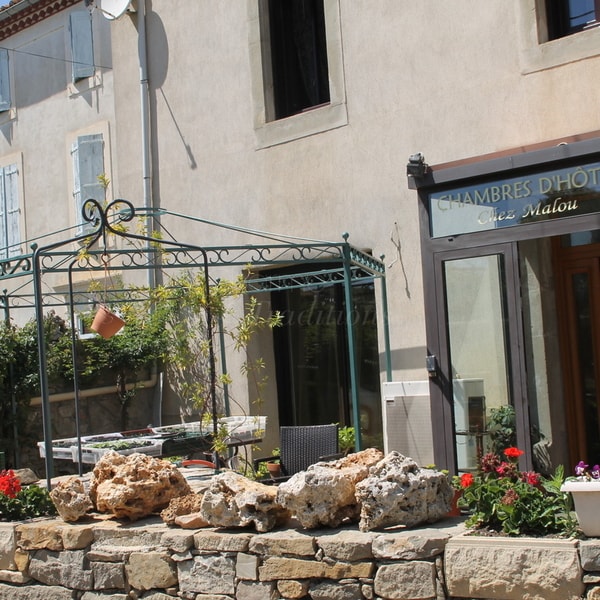 A charming outdoor seating area features a decorative wrought-iron gazebo surrounded by vibrant flowers. Natural stone walls underline the space, complemented by a table and chairs. A nearby building showcases glass accents, indicating a welcoming entrance for guests.