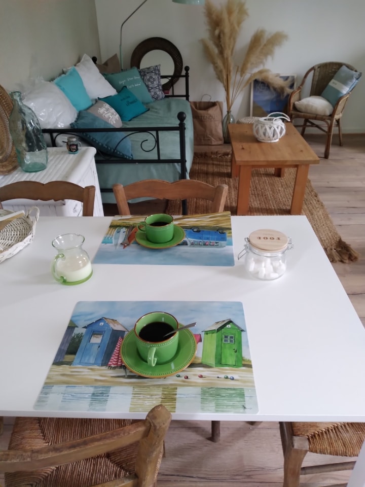 A dining table is set with green cups and a coffee pot on vibrant placemats featuring coastal artwork. Nearby, a cozy seating area is visible with a light green sofa adorned with decorative pillows. Natural elements like pampas grass enhance the inviting space.