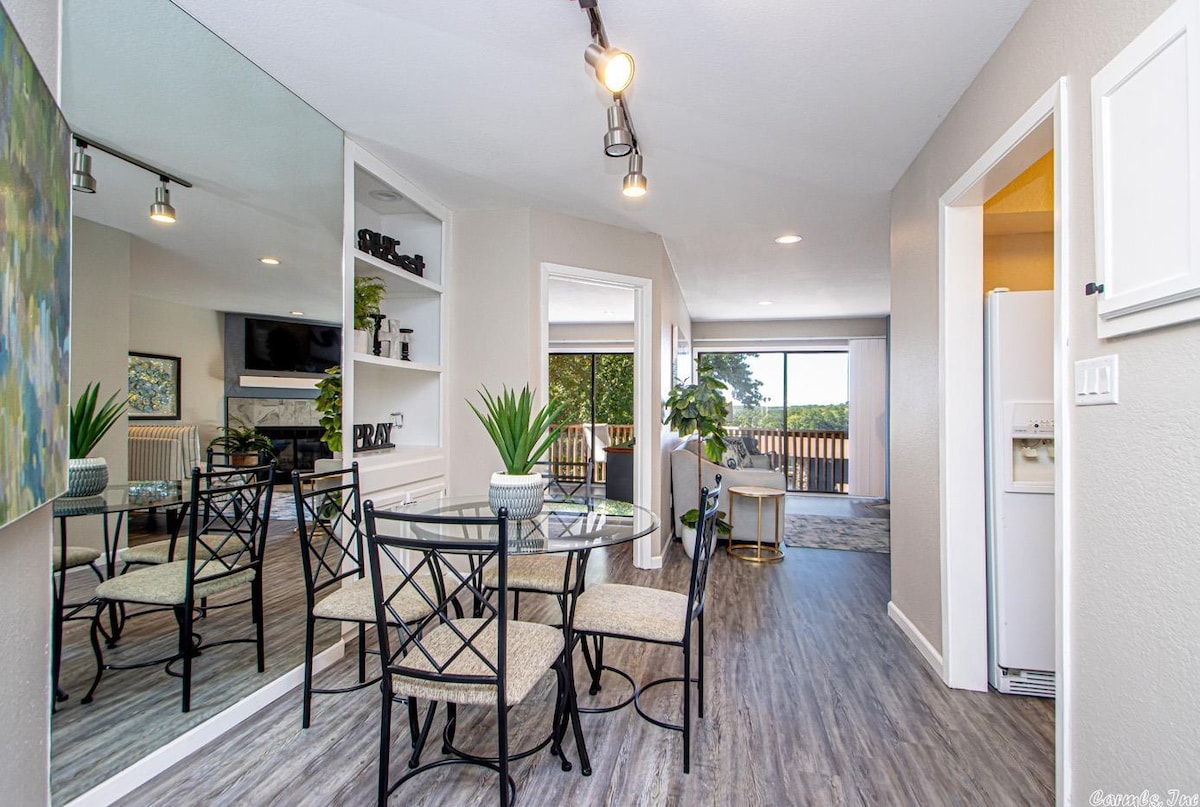 A dining area features a glass table surrounded by four black chairs, positioned near a large mirror. Natural light flows in from the nearby glass door, which leads to a balcony with a view of the outdoors. The open layout enhances the airy feel of the space.