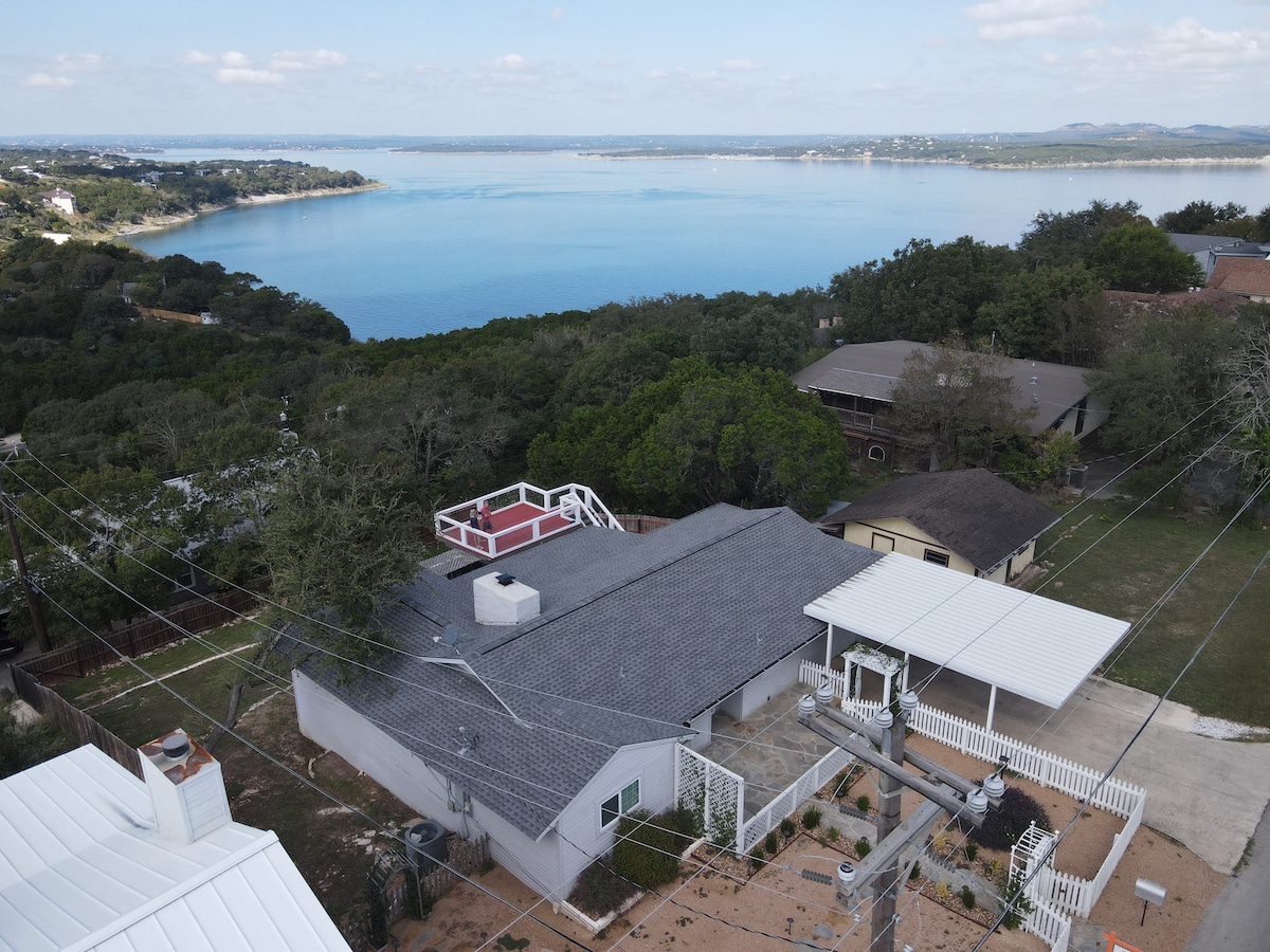 An aerial view showcases the Lake View Retreat nestled among lush greenery, with expansive water views of Canyon Lake in the background. The home's roof and outdoor spaces are visible, offering a glimpse of the serene surroundings and well-maintained landscape.