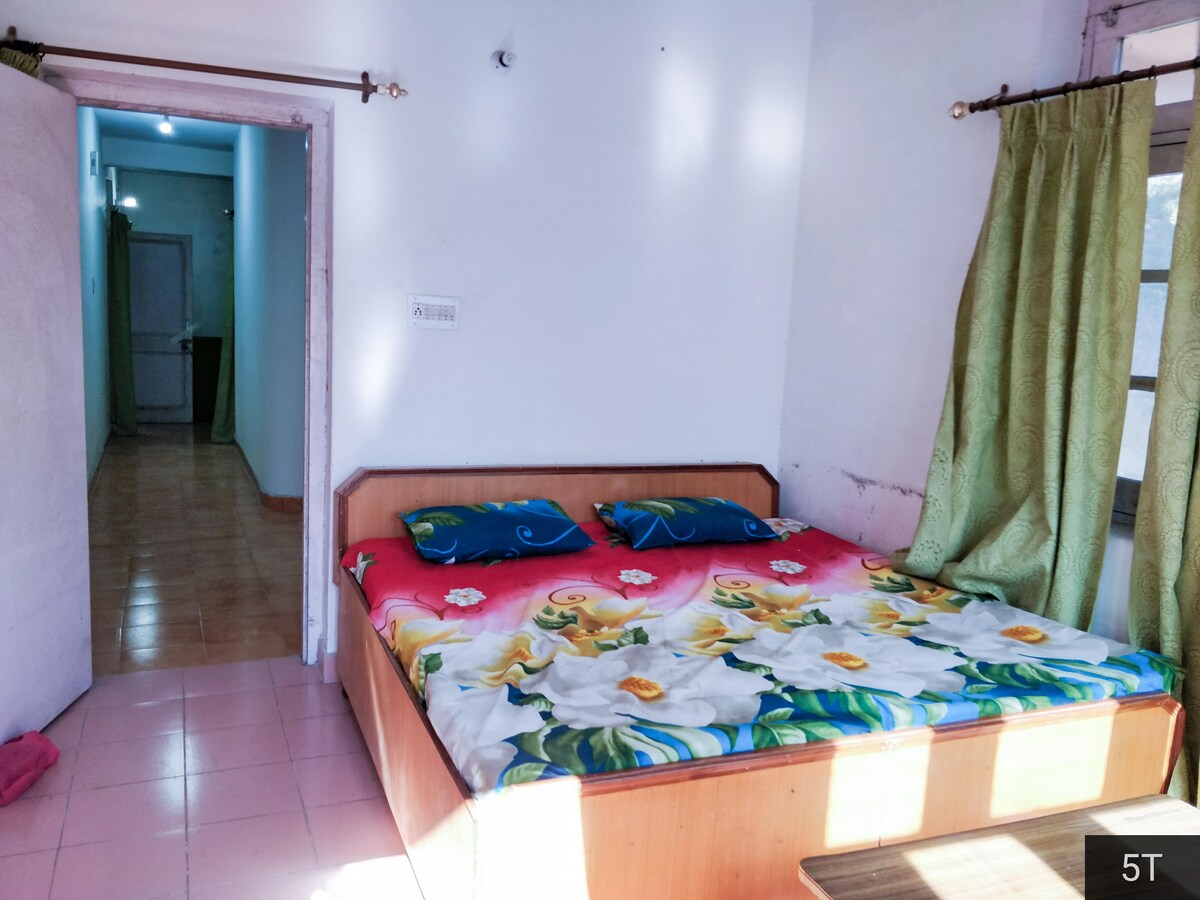 A spacious bedroom features a wooden bed with a floral-patterned bedspread. Light green curtains frame the window, and a small table is positioned nearby. A doorway leads to another area, providing a glimpse of the homeās layout.