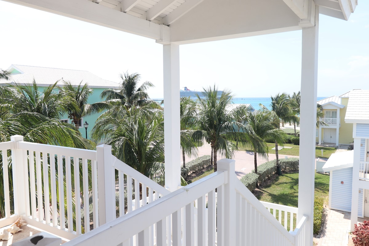 A view from a balcony presents swaying palm trees and a pathway leading towards the beach. The ocean glistens in the distance, visible beyond lush greenery and nearby buildings. Sunlight brightens the scene, creating a relaxed and inviting outdoor setting.