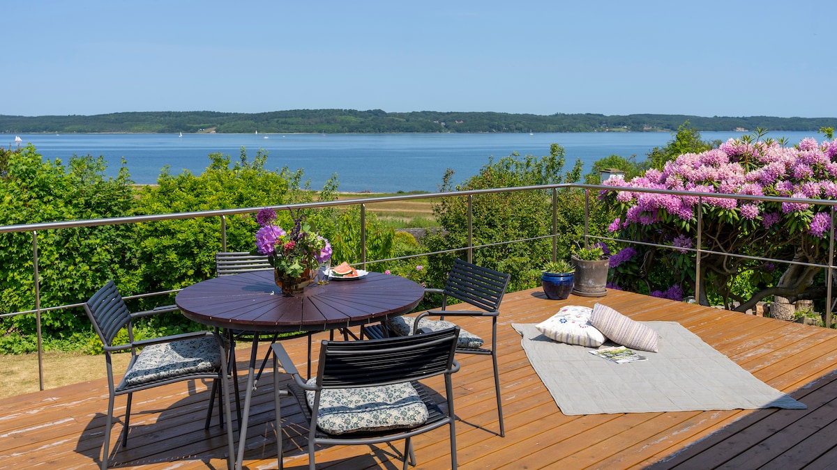 An outdoor terrace offers a round wooden table surrounded by four black chairs, all positioned to take advantage of the breathtaking view over Vejle Fjord. Lush greenery and blooming rhododendrons enhance the setting, creating a serene spot for relaxation.