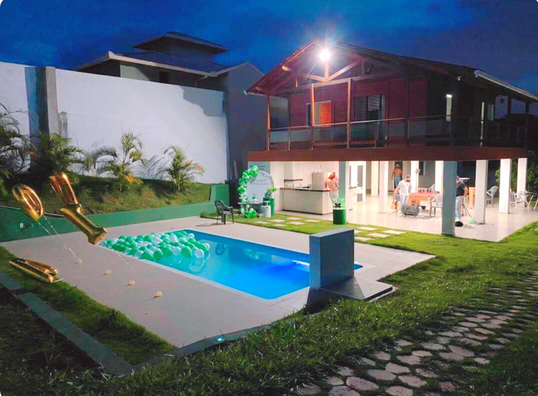 An outdoor area is showcased, featuring a swimming pool surrounded by green grass and decorative balloons. The two-story building has an open patio with seating and a kitchenette, inviting gatherings. Soft evening lighting enhances the relaxed atmosphere.