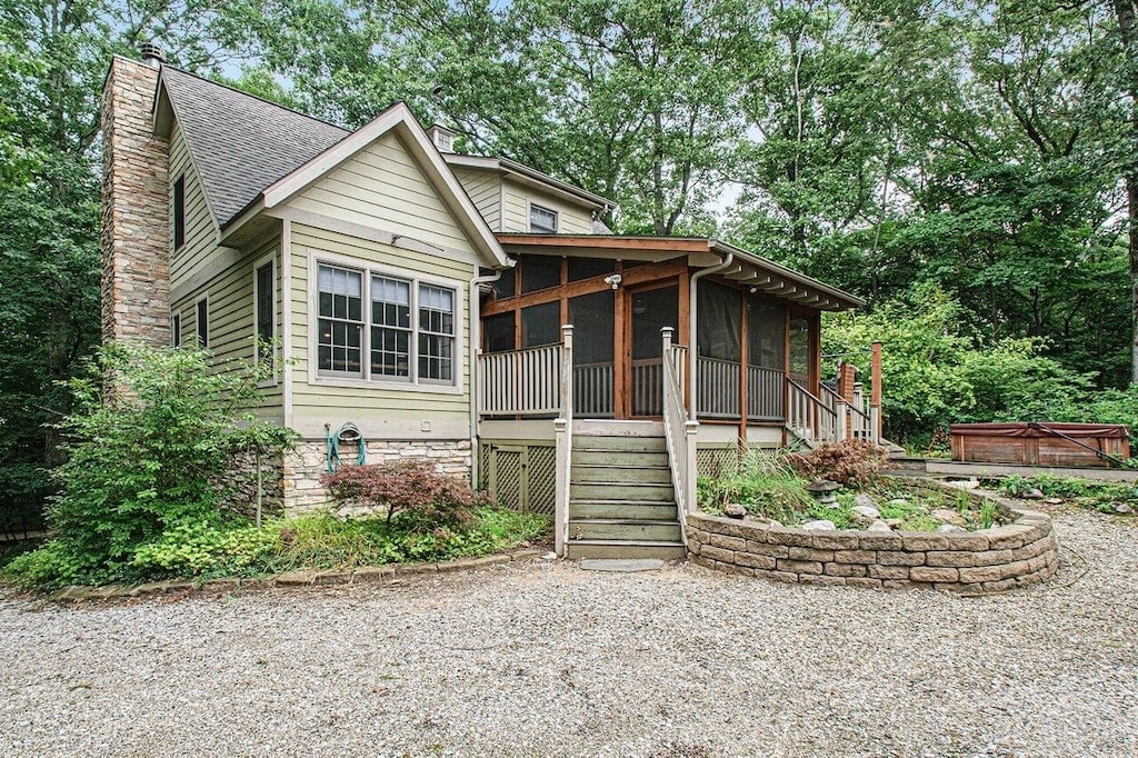 The lodge exterior features a combination of wooden and stone finishes, surrounded by lush greenery. A welcoming approach is created by the gravel driveway leading to a set of steps leading to a screened porch, which offers a view of the forested surroundings.