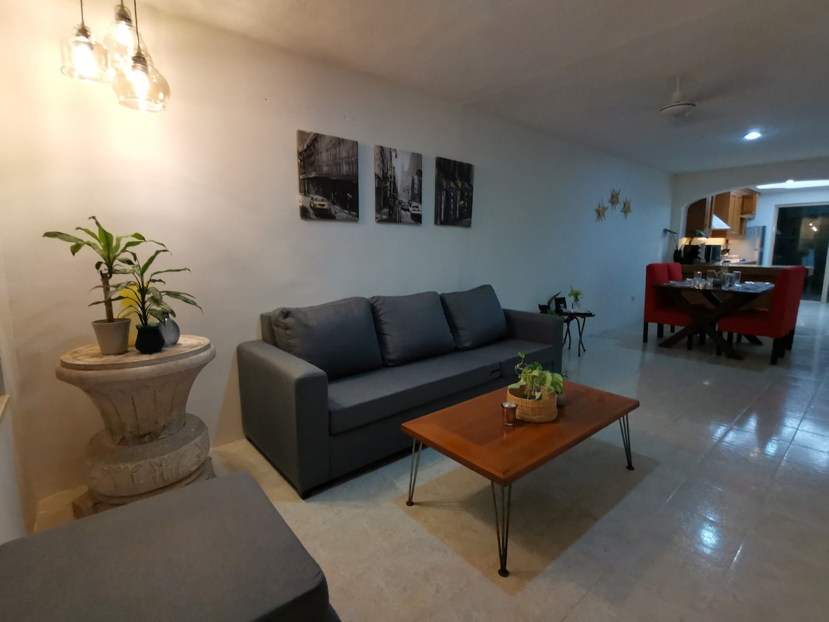A spacious living area features a gray sofa paired with a wooden coffee table, creating a comfortable seating arrangement. Wall art depicting urban scenes adds visual interest. Soft lighting is provided by pendant fixtures, illuminating the space, which leads to the kitchen and dining area.